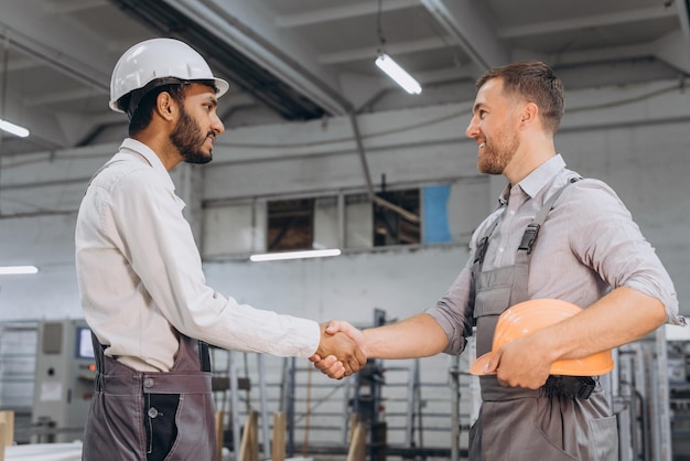 Two international male workers in overalls shake hands against the background of the production of PVC windows and doors