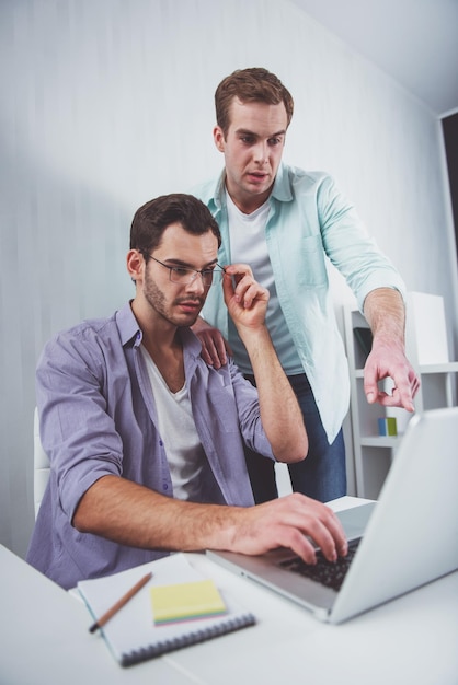 Two young attractive businessmen in casual clothes using laptop and discussing while working in office