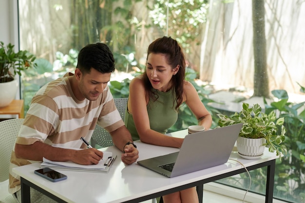 Two young intercultural colleagues organizing work or analyzing online data while man making notes on paperwork during discussion