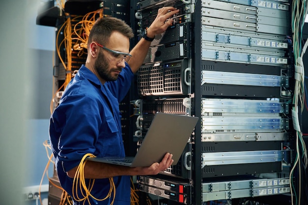 Using the laptop Young man is working with internet equipment and wires in server room