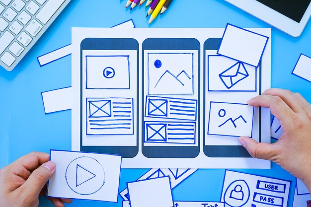 Working desk with hands sorting wireframe screens of mobile responsive website Developing wireframe sketch layout design mockup on smartphonetablet screen