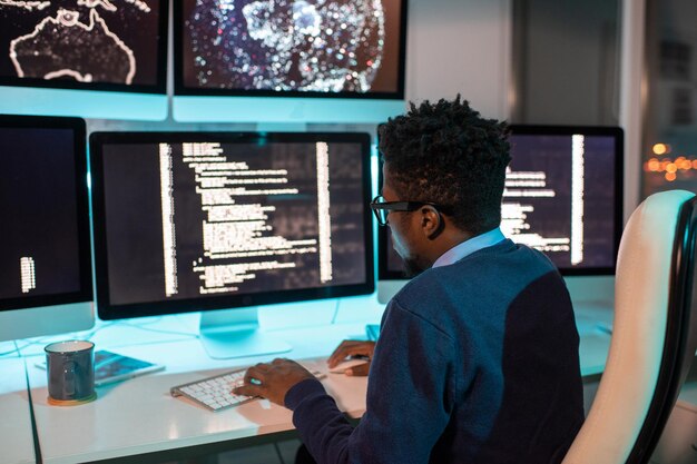 Young African male software developer working late in front of computer screen with coded data in office environment