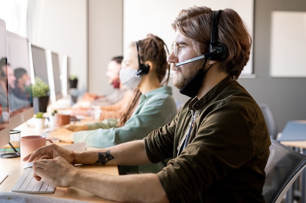 Young serious bearded male operator with protective mask under chin consulting clients while sitting in front of colleagues in headsets