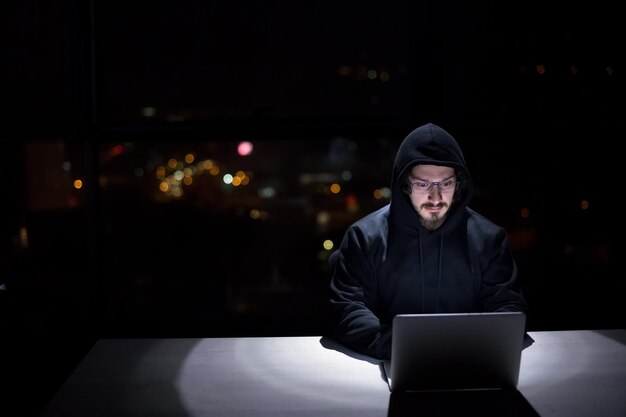 Young talented hacker using laptop computer while working in dark office with big city lights in the background at night