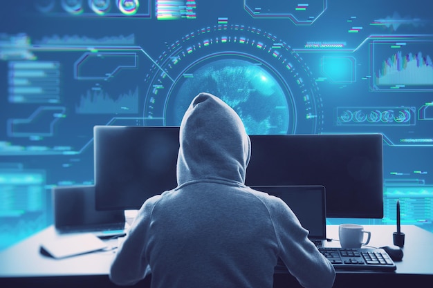 back view of hacker in hoodie sitting at desktop with coffee cup and using computer monitors