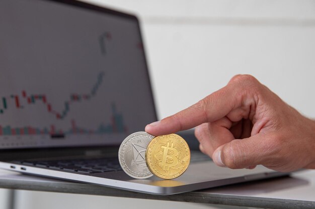 Close-up of Bitcoin and Ethereum cryptocurrencies held up by a finger with candlestick charts in the background of a laptop. Trading of cryptos, new virtual money. Crypto currency.