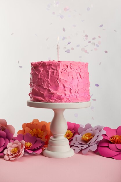 Delicious pink birthday cake with candle on cake stand near paper flowers and confetti on grey