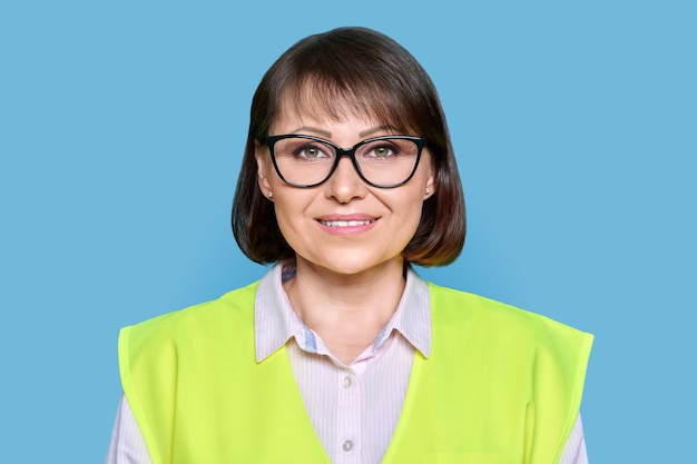 Headshot of industrial mature woman in protective vest glasses on blue isolated background