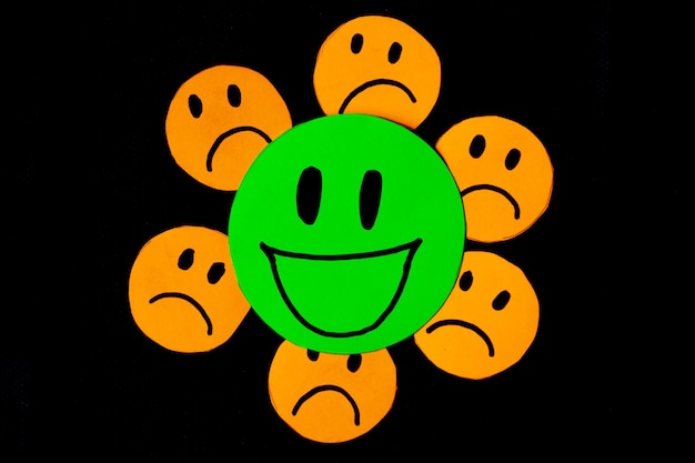 image of some emoticons cut out of orange paper with a sad face surrounding a big green emoticon with a smiling face concept of bad influences bad friendships negativity problems bullying