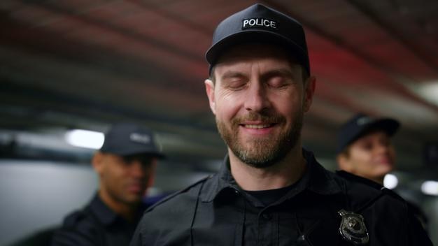 Kameraya gülümseyen polis memurunun portresi. Şapka ve polis üniforması giyen erkek polis memuru. Kameraya poz veren pozitif polis memuru. Devriye arabasının yanıp sönen ışıklarıyla arka planda duran meslektaşlar.