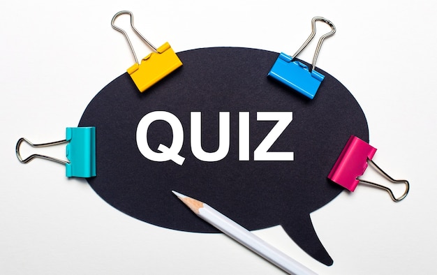 On a light surface, multi-colored paper clips, a white pencil and black paper with the words QUIZ