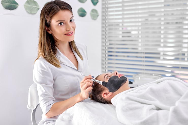 Fotoğraf people, beauty, spa, cosmetology and skincare concept. beautiful young woman at spa lying in white bathrobe, professional nice and pleasant confident cosmetologist applying facial mask using brush