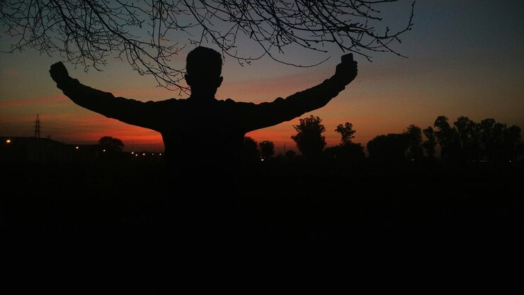 Fotoğraf rear view of man with arms outstretched standing against sky at sunset