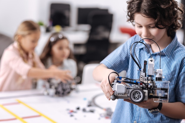 Representing my invention. Cheerful gifted inspired boy standing at school and holding robot while his classmates working on the project