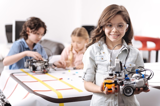 Showing my talent. Smiling cheerful happy girl standing at school and holding robot while colleagues working on the project