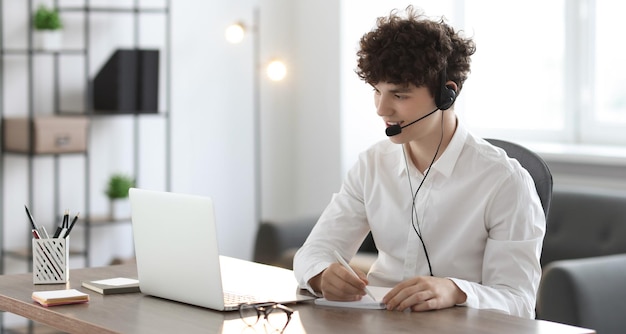 Teenager in headset taking notes while working with laptop at table indoors Remote job
