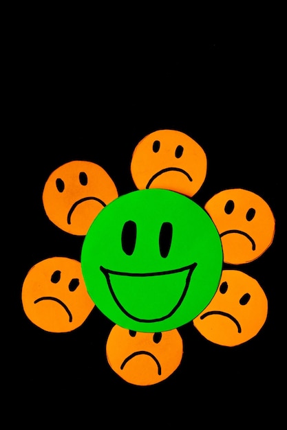 vertical image of some emoticons cut out of orange paper with a sad face surrounding a big green emoticon with a smiling face concept of bad influences bad friendships negativity problems bullying