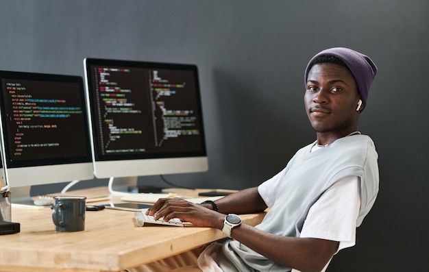 Young black man in casualwear keeping hands on computer keyboard while sitting in front of monitors
