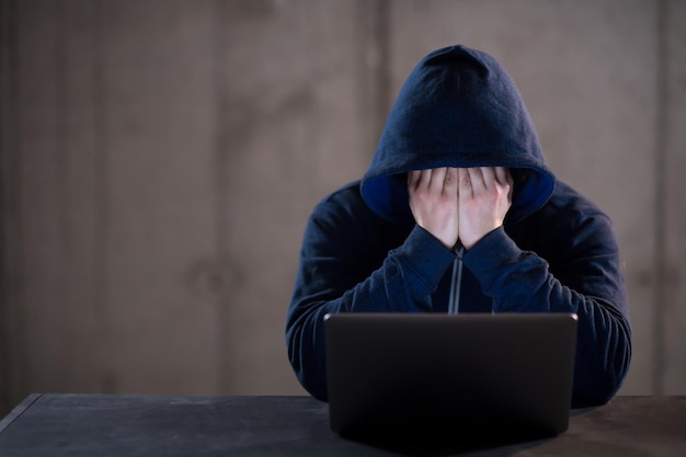 Young talented hacker using laptop computer while working in dark office with concrete wall in the background
