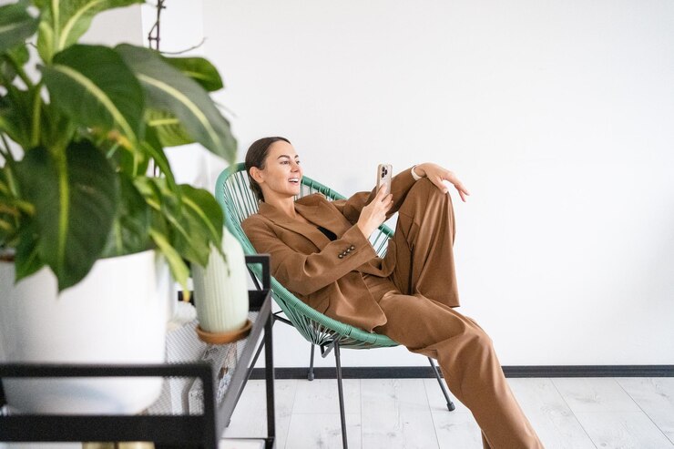 Fotoğraf young woman using phone and laughing at camera she is wearing stylish suit and seating in chair