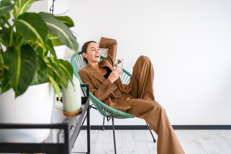 Fotoğraf young woman using phone and laughing at camera she is wearing stylish suit and seating in chair