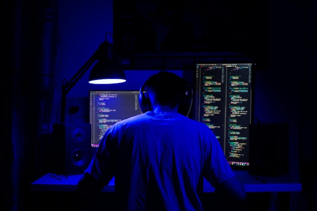 A man sits at a computer in a room at a table at night with blue lighting and programs