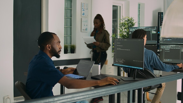 African american developer using laptop to write code and display system information on computer. Database coder parsing algorithm working on user interface in software agency.