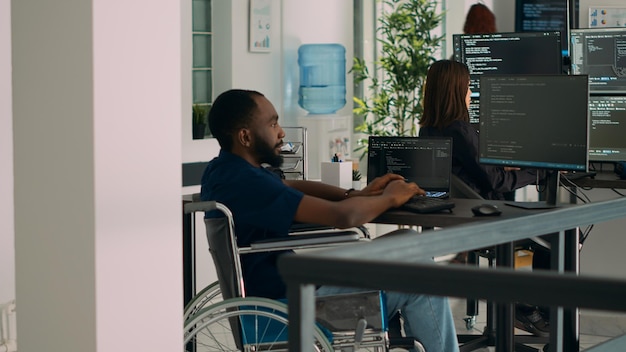 African american IT engineer with disability typing code in it development agency office, using terminal window to write html script database. Wheelchair user working with security programming.