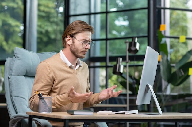An angry young man sits at the table looks at the computer screen spreads his hands does not