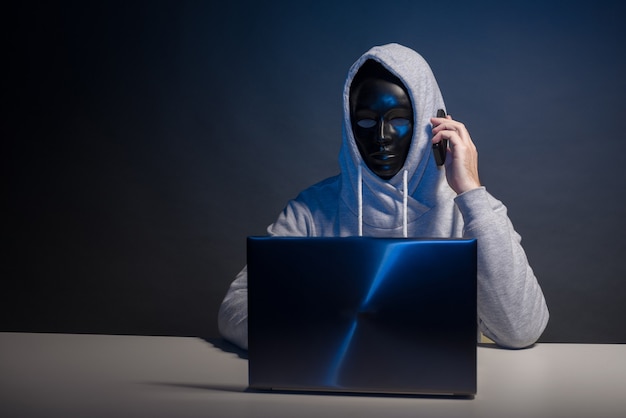 Anonymous hacker in mask programmer uses a laptop and talking on the phone to hack the system in the dark. The concept of cybercrime and hacking database