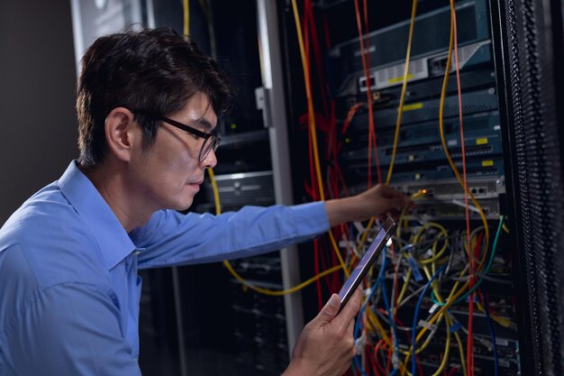 asian male engineer using digital tablet and inspecting computer server in computer server room. database server management and maintenance concept