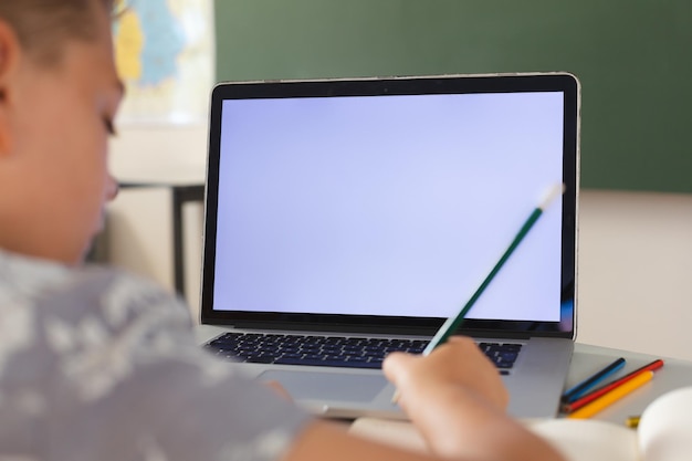 Caucasian boy sitting at a desk in classroom writing and using laptop, with copy space on screen