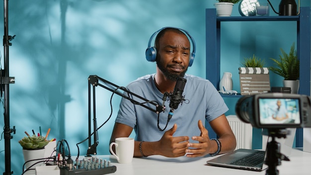 Cheerful vlogger waving at camera while recording podcast with microphone and headphones. Influencer filming vlog for social media channel using modern equipment and technology.