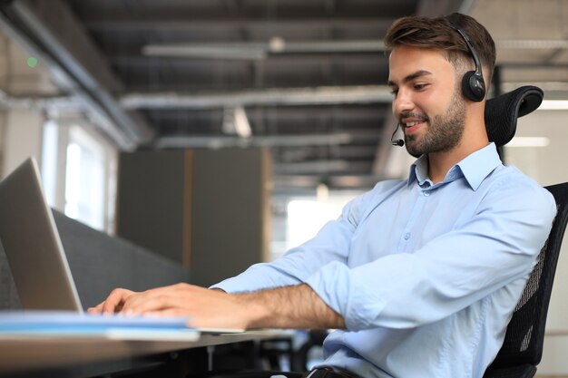 Cheerful young support phone male operator in headset, at workplace while using laptop, help service and client consulting call center concept.
