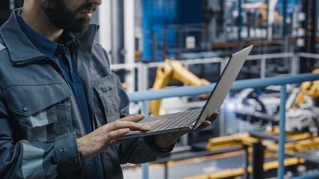 Close Up of Automotive Industry Engineer in Grey Uniform Using Laptop at Car Factory Facility Professional Assembly Plant Specialist Working on Manufacturing Modern Electric Vehicles