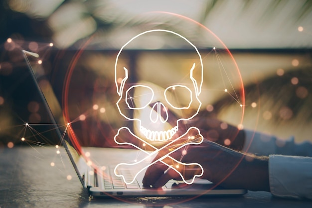 Close up of male hand using laptop and cellphone at desktop with digital red skull on blurry background Ransomeware virus and pirate threat concept Double exposure