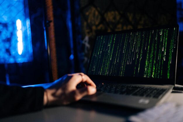 Closeup of unrecognizable hacker engaged in hacking into security system on laptop green binary code on screen with sitting at desk in dark basement room