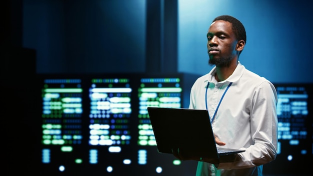 Computer scientist in high tech facility using laptop to patch network issues affecting server cabinets performance and connectivity, leading to slow data transfer rates and poor client communication