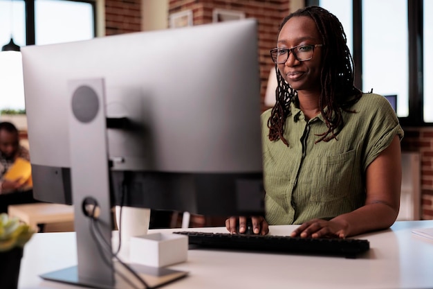 Confident african american freelancer looking at computer screen and typing while working remote from home. Focused programmer coding using pc in home living room while roommate is relaxing.