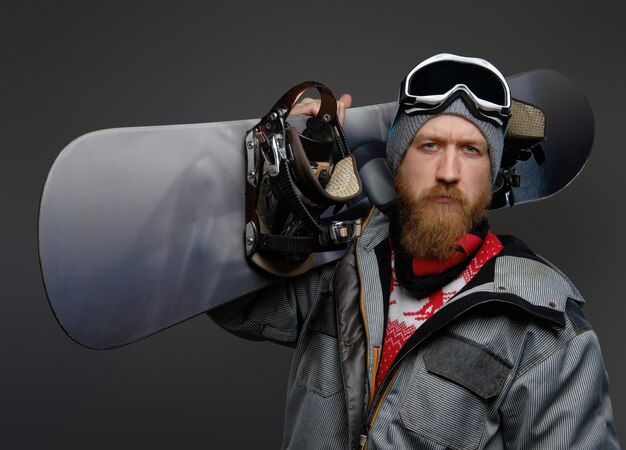 Confident man with a red beard wearing a full equipment holding a snowboard on his shoulder, looking at a camera with a serious look