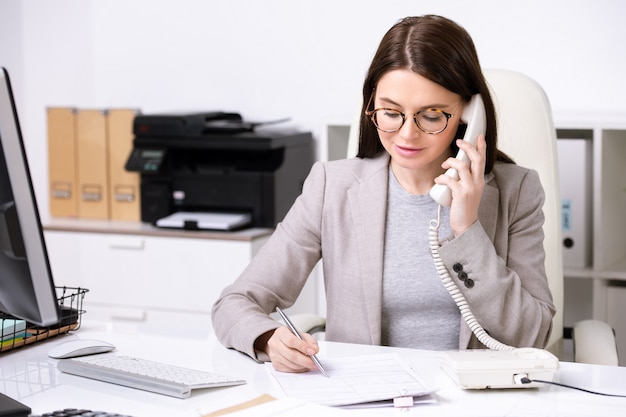 Confident young businesswoman sitting at table and making notes in document while discussing contract content by phone