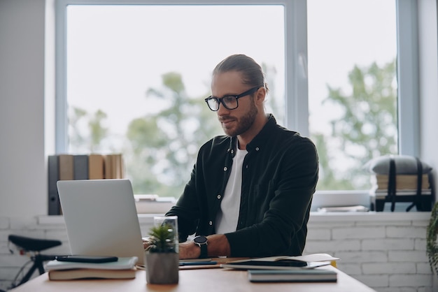 Confident young man using laptop while sitting at his working place in office