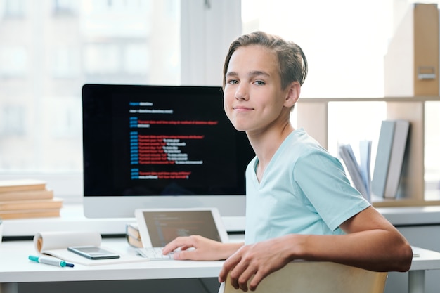contemporary boy in casualwear looking at you while sitting by desk with computer monitor in front at school