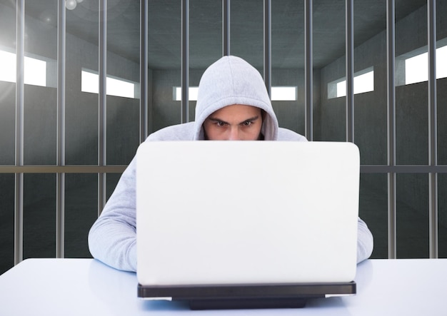 Criminal in hood with laptop behind prison bars