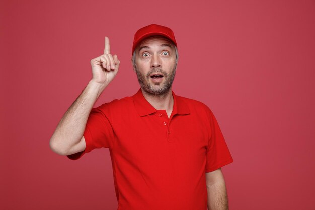 delivery man employee in red cap blank tshirt uniform looking at camera surprised showing index finger having great idea standing over red background