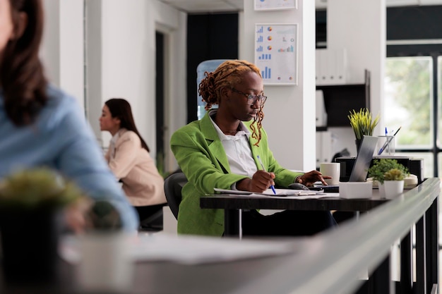 Employee working on laptop with financial report in business office open space, woman sitting at workplace desk. Young woman thinking, looking at documents side view medium shot