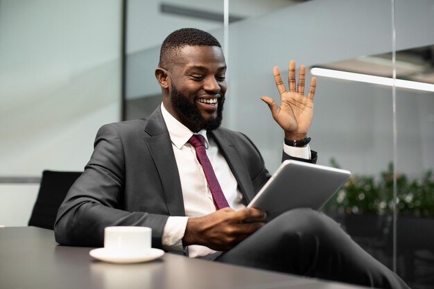 Happy black businessman in expensive suit sitting at table in office having video chat with business partners using digital tablet looking at gadget screen smiling and gesturing copy space