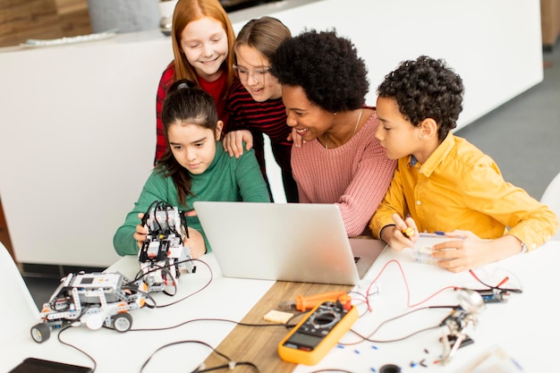 Happy kids with their African American female science teacher with laptop programming electric toys and robots at robotics classroom