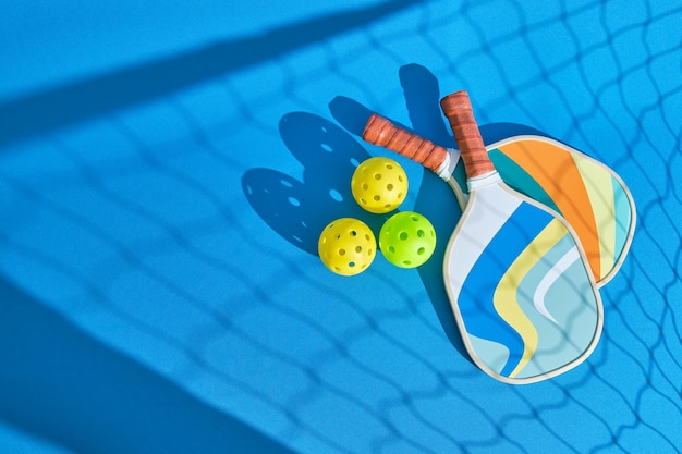 image from above of two pickleball paddles on the blue floor with the shadow of the net in front and some balls to the side