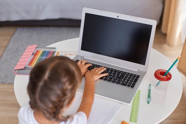 Indoor shot of dark haired female child with braids typing on keyboard doing her school tasks distance education lessons at home little girl sitting at table and studying on laptop